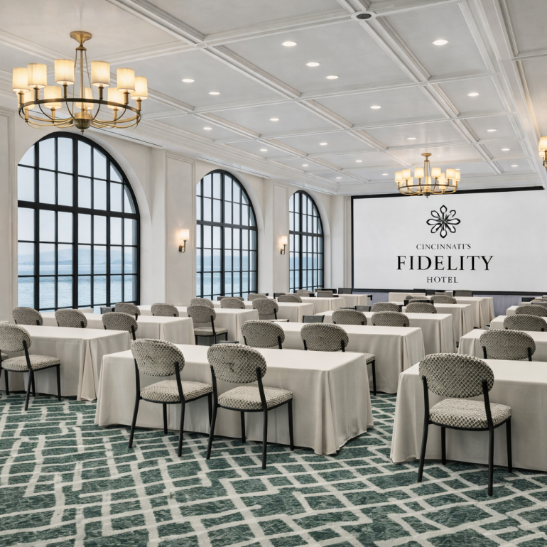 A large conference room displays rows of tables and patterned chairs facing a screen with:
CINCINNATI'S
FIDELITY
HOTEL
Arched windows overlook water, with chandeliers illuminating the space.
