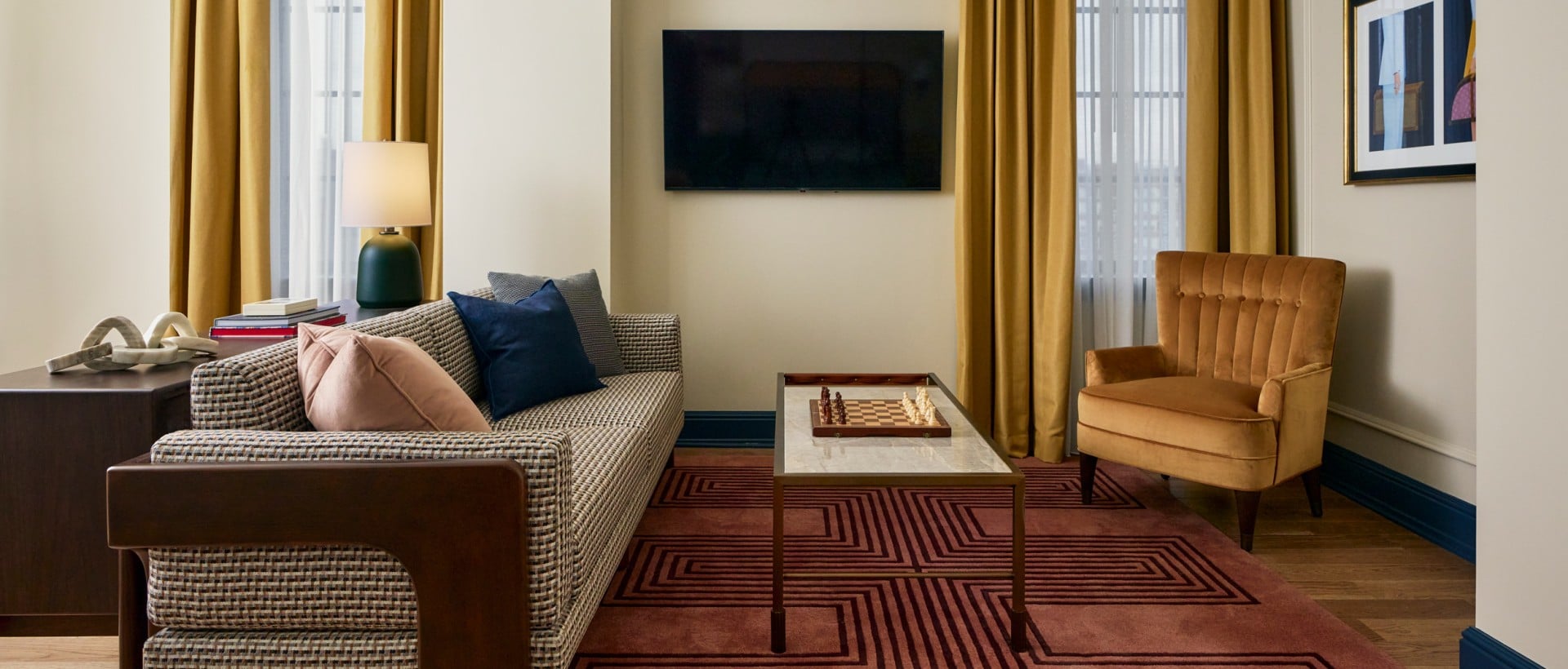 A patterned sofa, marble coffee table with a chess set, and gold armchair fill a modern living room. Gold curtains, a geometric rug, and wall-mounted TV complete the space.