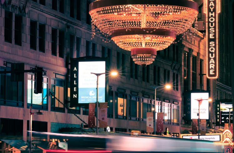 A large, tiered crystal chandelier illuminates a city street at night. Blurred vehicle lights streak past buildings with numerous glowing signs.
