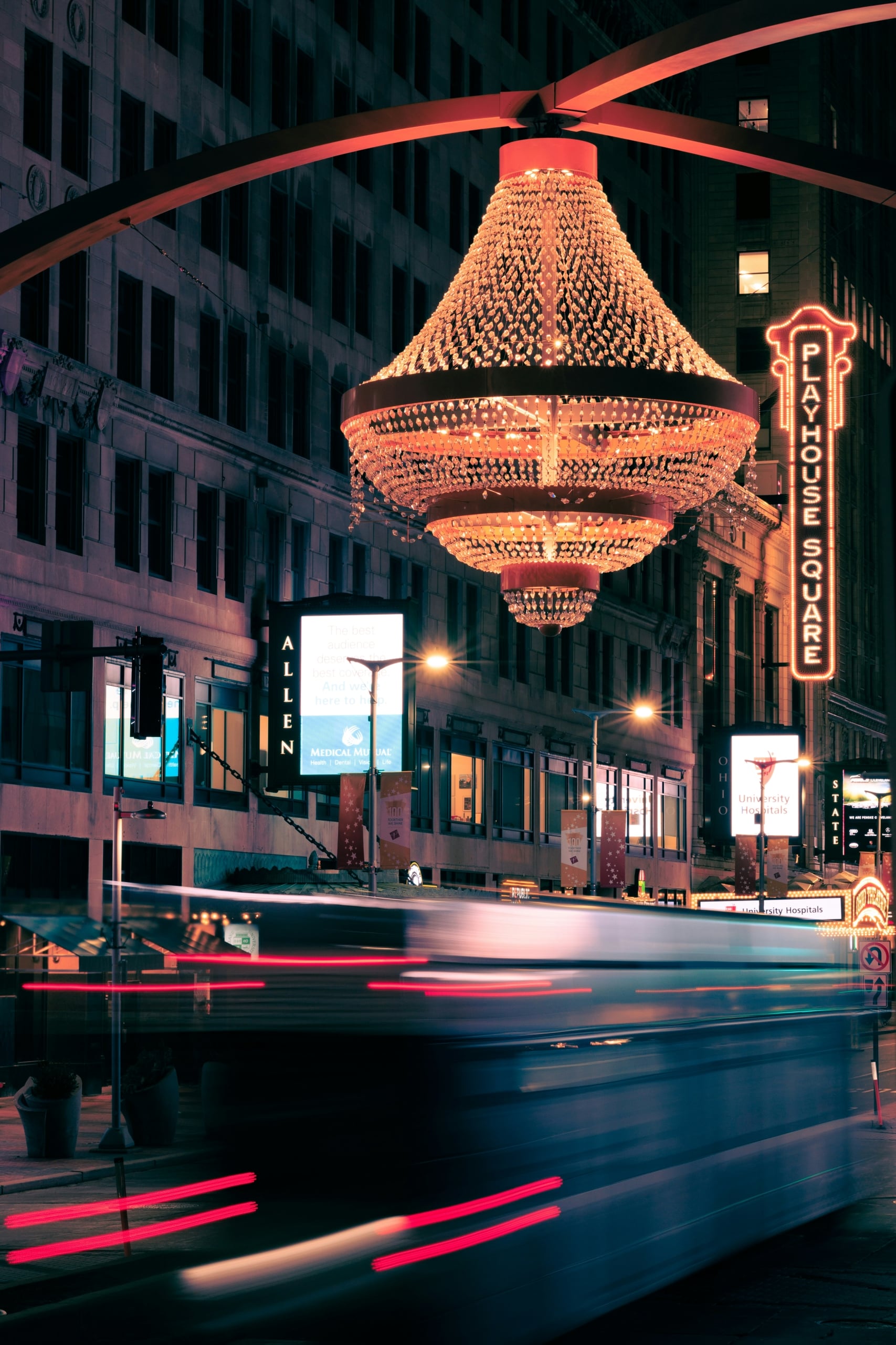 A large, tiered crystal chandelier illuminates a city street at night. Blurred vehicle lights streak past buildings with numerous glowing signs.