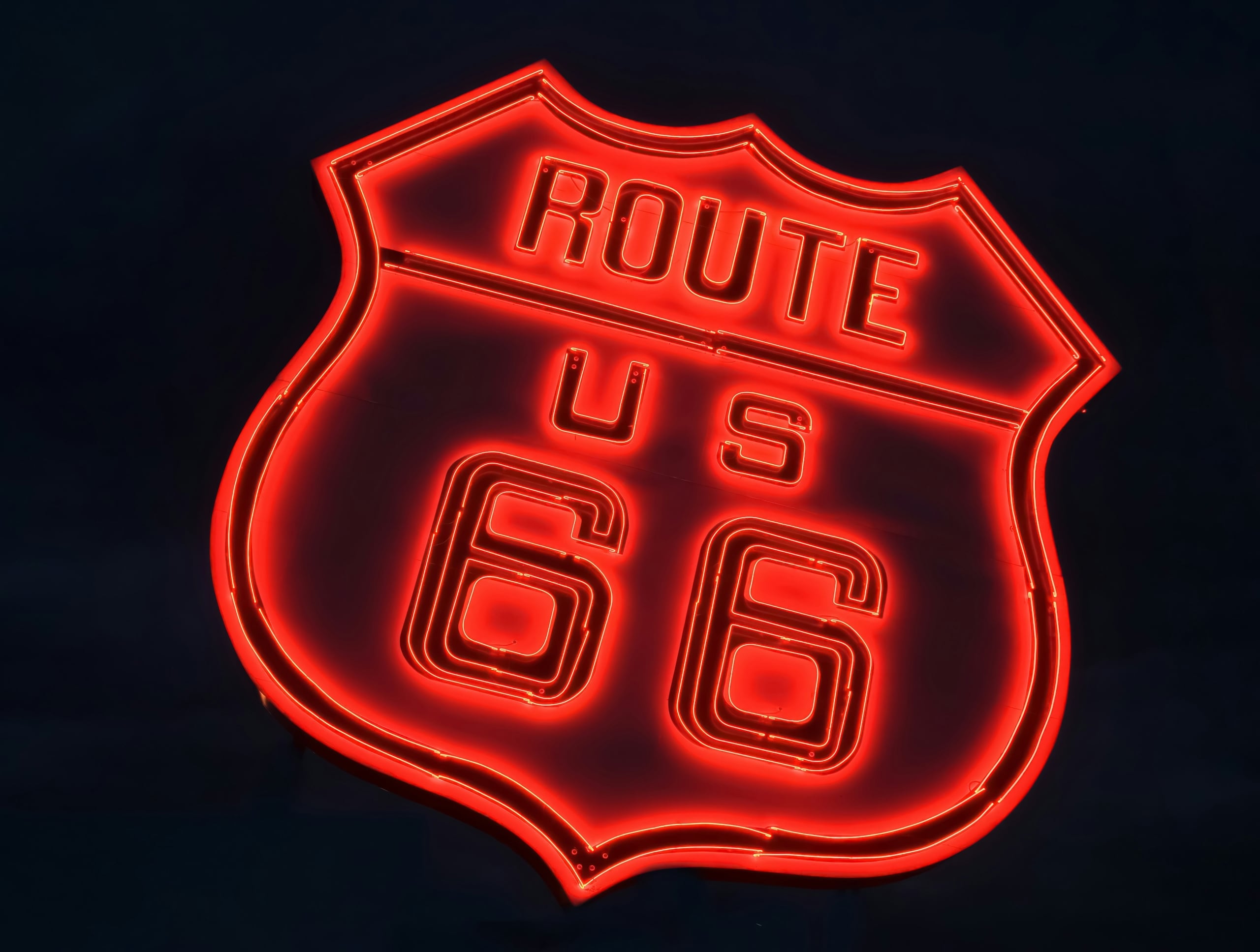 A red neon sign in the shape of a U.S. highway shield glows brightly against a dark background, displaying the text: ROUTE U S 66.