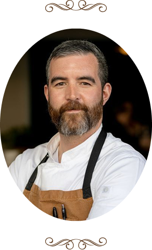 A bearded man looks forward, wearing a white chef's jacket and brown apron with pens, framed in an oval against a dark background, with decorative scrolls above and below.