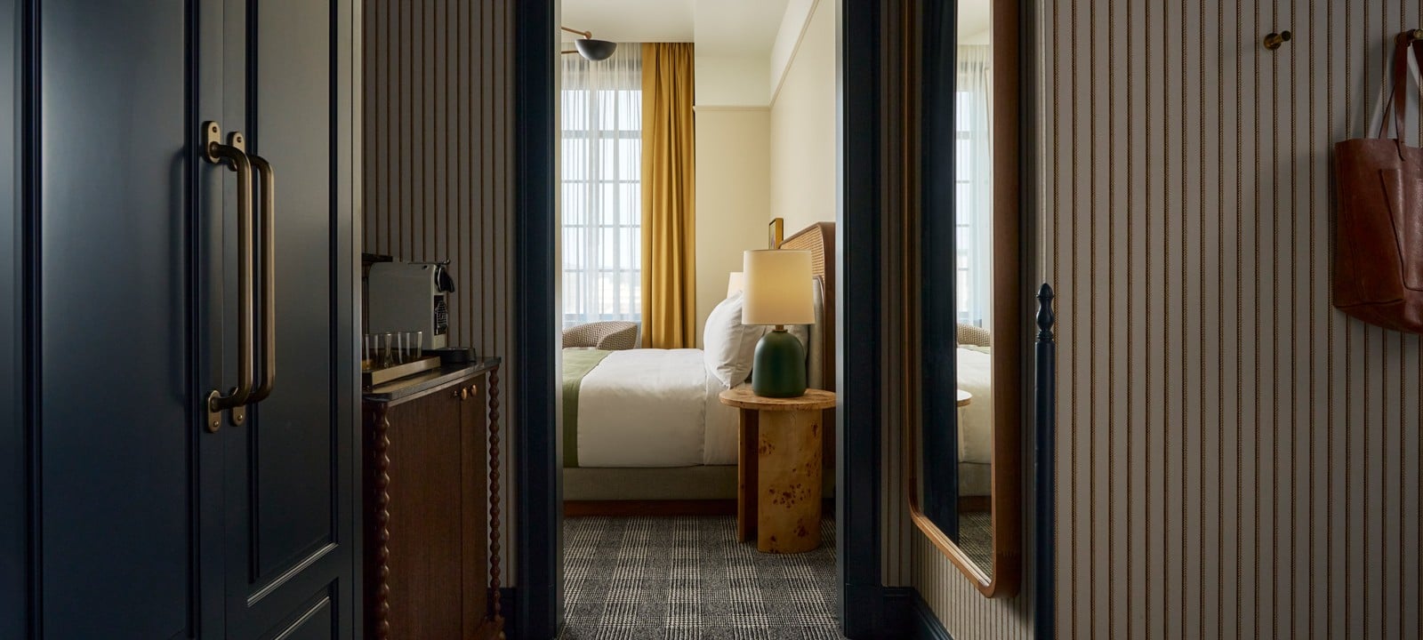 A dark doorway reveals a bedroom with a bed, lamp, and yellow curtains. The hall features patterned carpet, striped walls, a coffee machine, and a hanging leather bag.