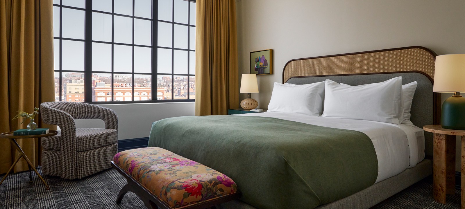 A modern hotel room contains a bed with green blanket and white pillows, facing a large grid-paned window revealing a city view. An armchair, floral bench, and plaid carpet are present.