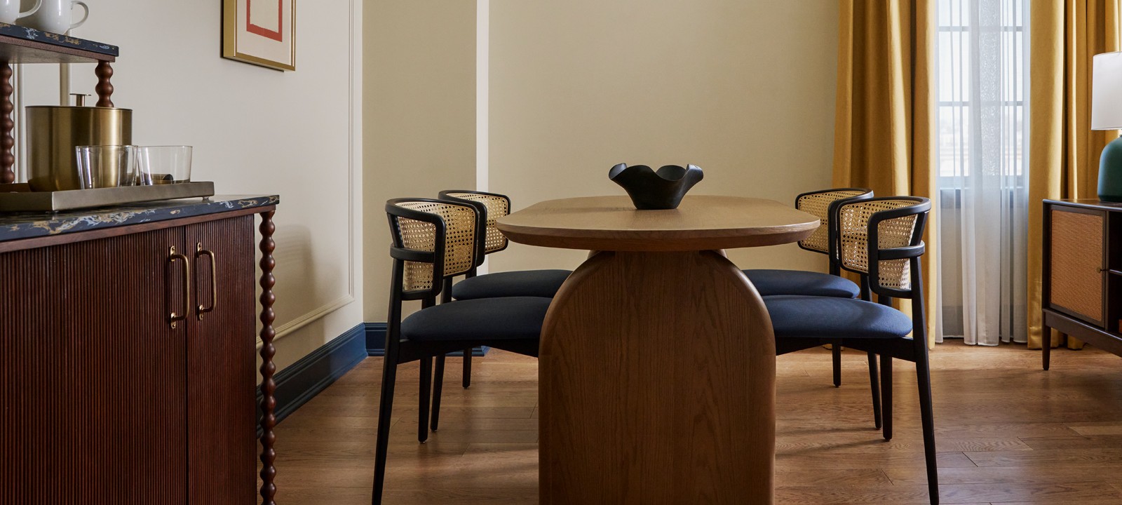 A wooden dining table with a black bowl is surrounded by four chairs with cane backs and blue cushions in a room with light walls, wood floors, and yellow curtains.