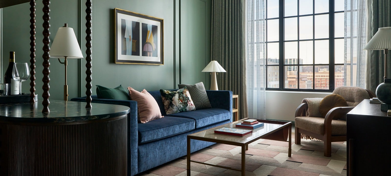 A blue velvet sofa with throw pillows fills a modern living room featuring dark green paneled walls, a large gridded window, a geometric rug, and a carved wooden bar.