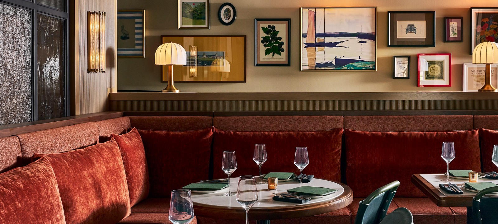 Restaurant dining area arranged for guests, with a long, reddish-brown velvet banquette, round tables set with dark green placemats, and a gallery wall displaying varied art. A frame reads: WINTON MOTORS 1905.