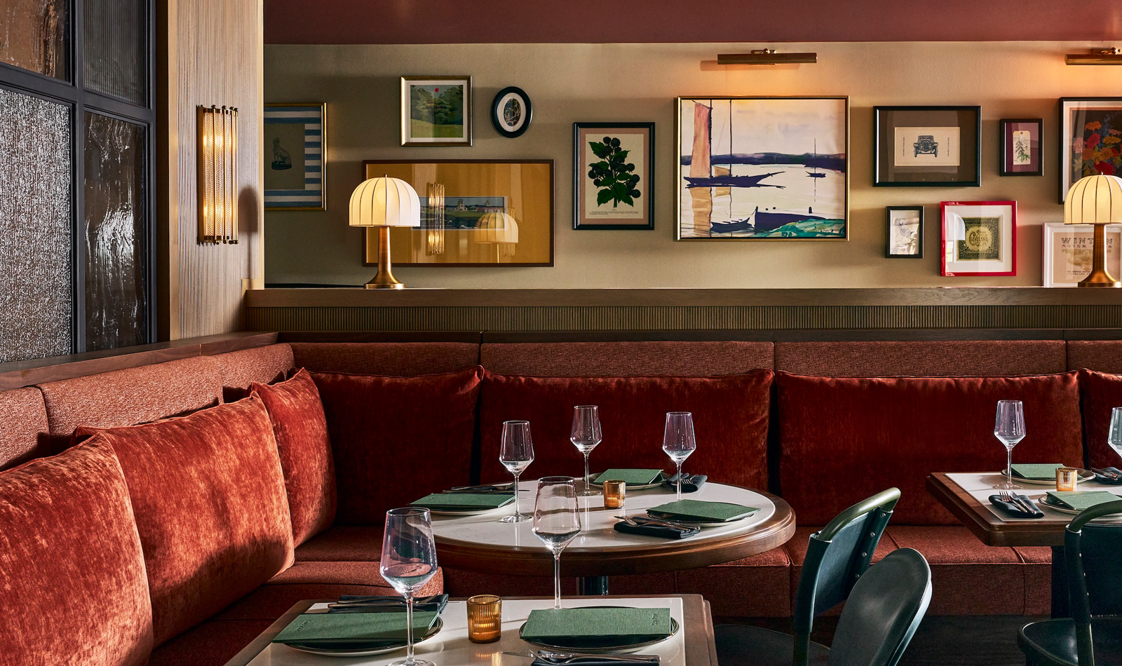 A dining area with a red velvet banquette, round tables set with green napkins and wine glasses, and framed art on the walls.