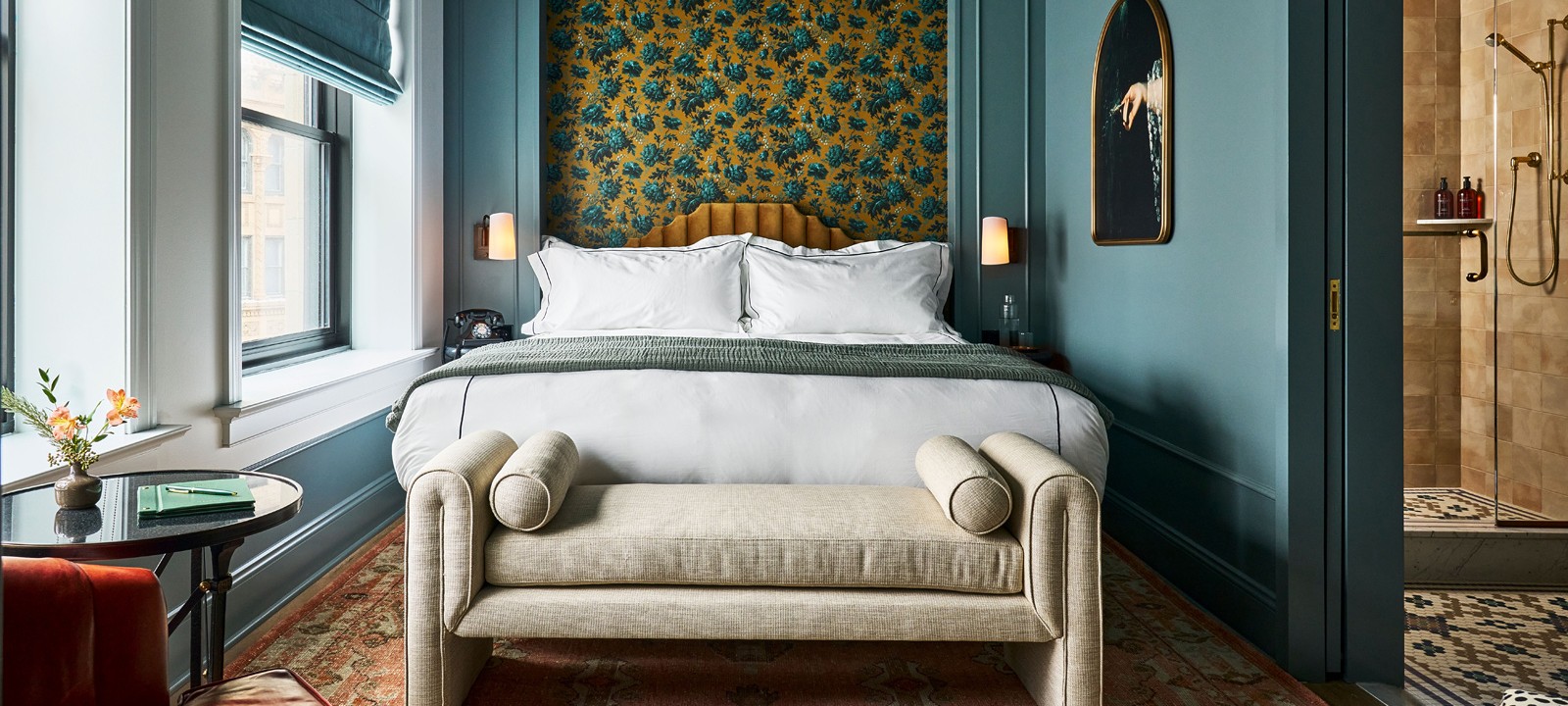 A modern bedroom features a large bed with a floral wallpaper headboard, a bench at its foot, teal walls, a window left, and a shower right.