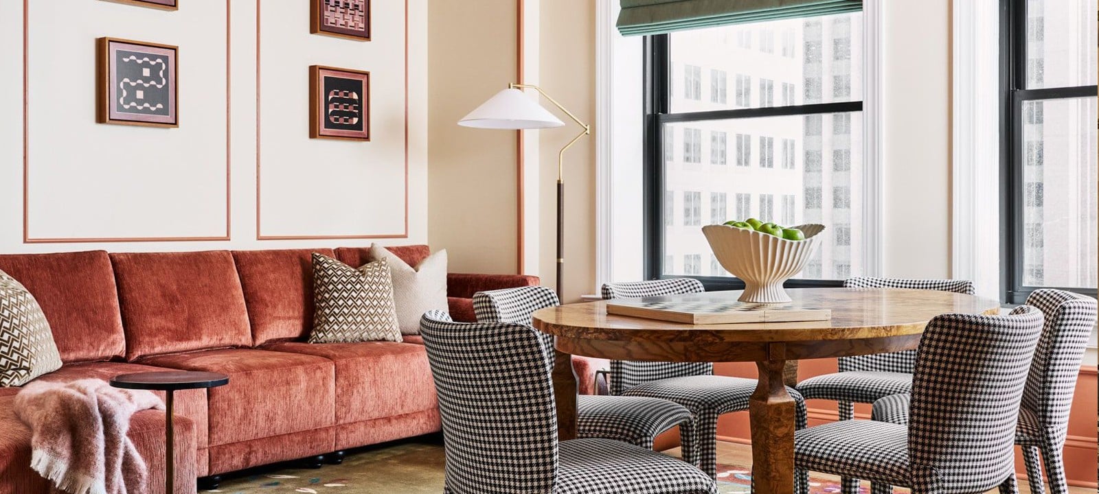 A modern living/dining room displays a long reddish-brown sofa, a round wooden table with houndstooth chairs, and wall art. Large windows overlook city buildings.