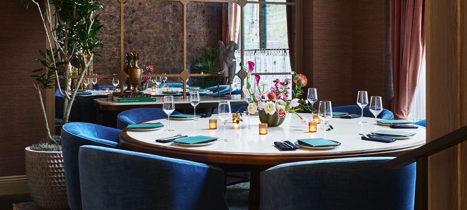 A large round dining table is elegantly set with blue velvet chairs, plates, glasses, and a floral centerpiece in a refined restaurant. Books: "A MAN & HIS CAR", "THE ARCHITECTURE OF TREES".