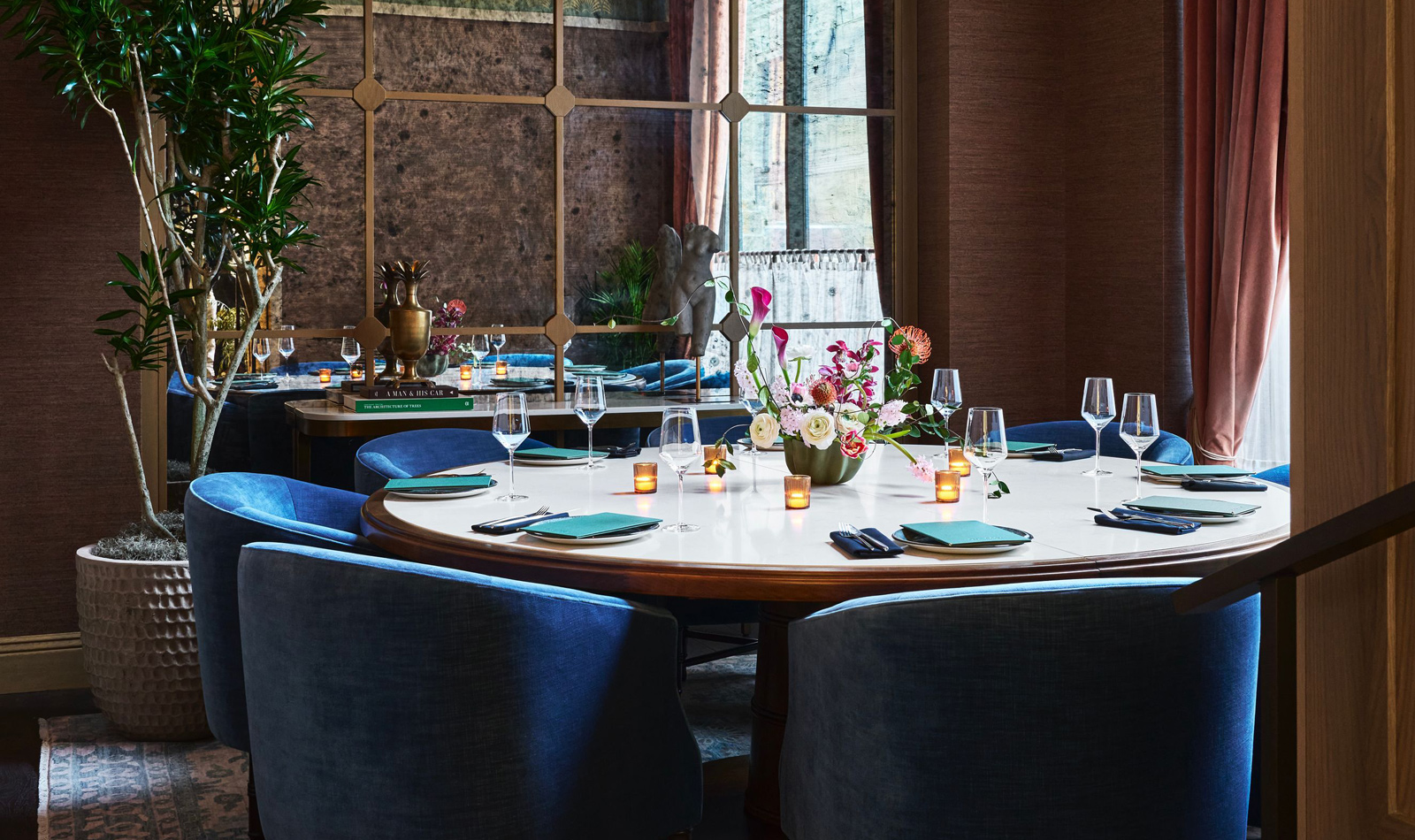 A round dining table set with blue chairs, wine glasses, and candles in a room with a large window.