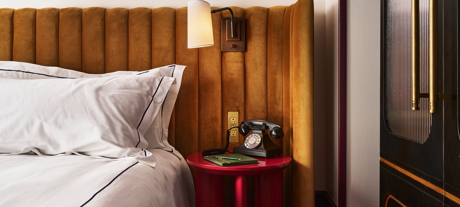 A mustard velvet headboard and white bedding flank a red bedside table holding a black rotary phone, green notebook, and pen, lit by a wall sconce.