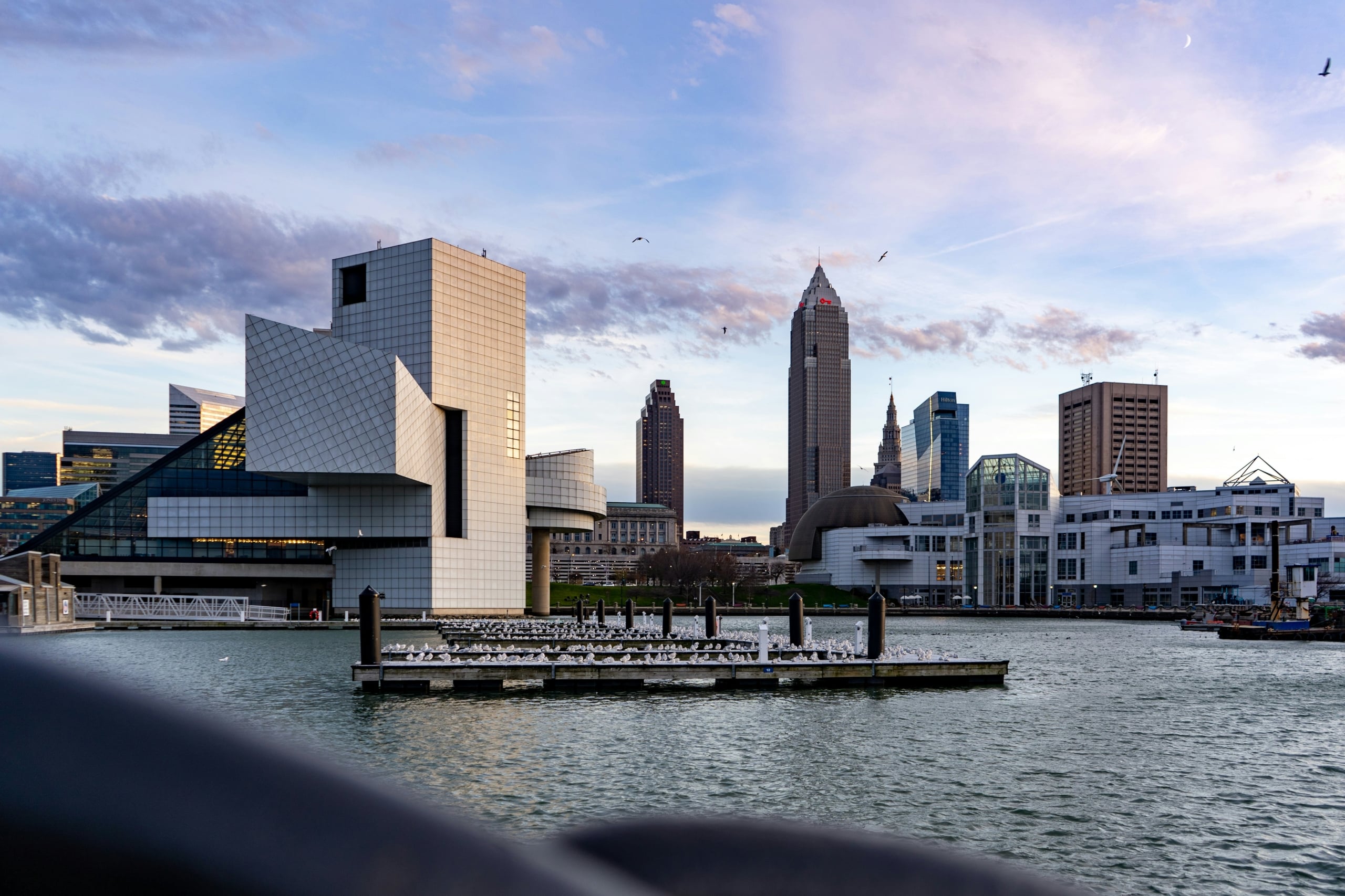 Angular Rock & Roll Hall of Fame building by a harbor. Seagulls cover a pier labeled "12". A city skyline, featuring "KEYBANK", "Hilton", and "Huntington" towers, frames the background under a cloudy sky.