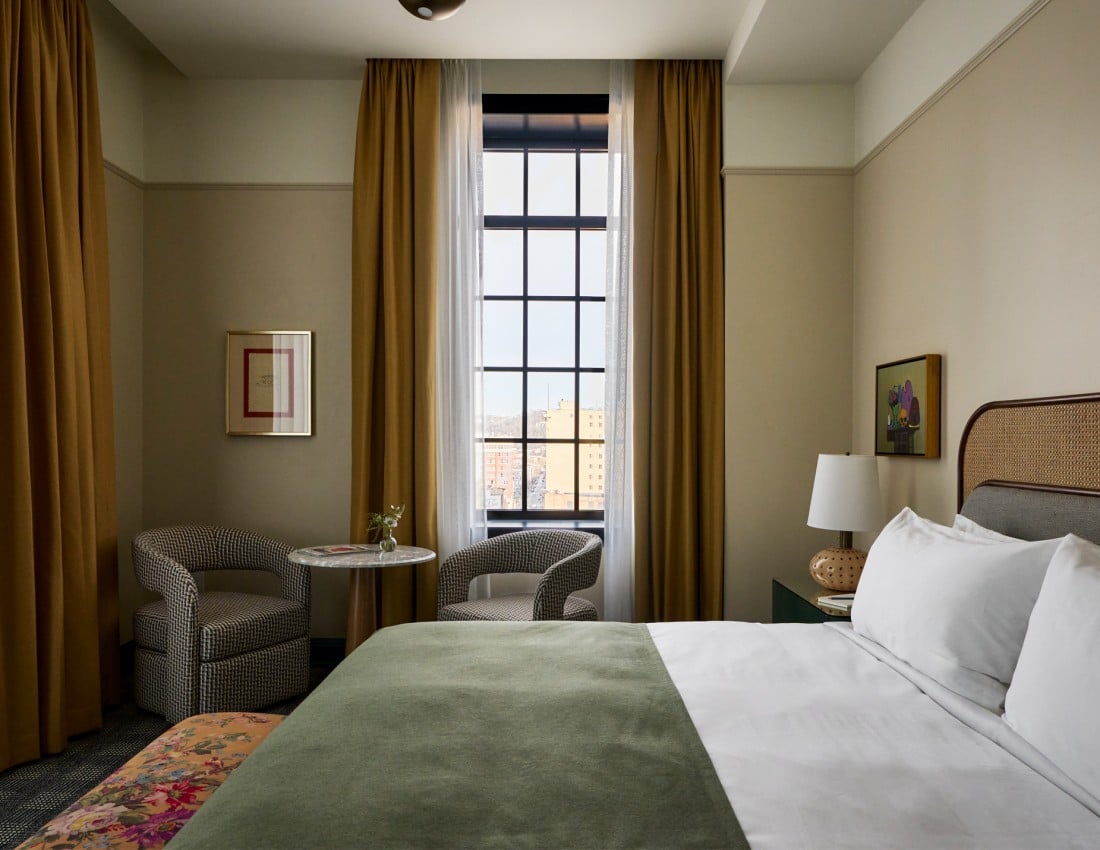 A neatly made bed with a green throw invites rest in a hotel room featuring a tall window with city views, two armchairs, and artwork on cream walls.