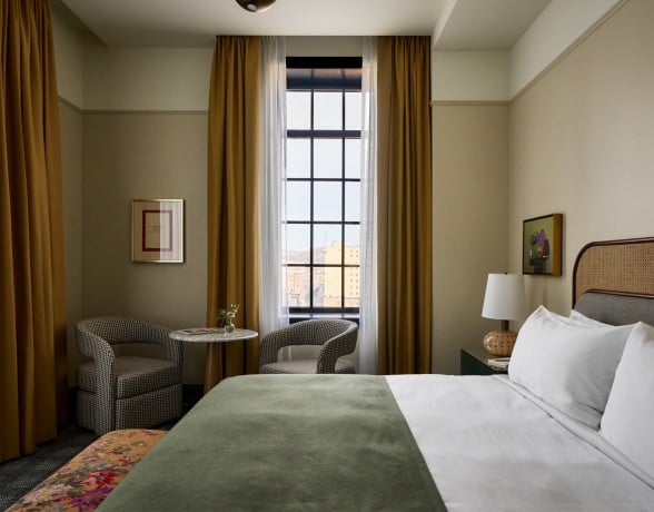 A neatly made bed with a green throw invites rest in a hotel room featuring a tall window with city views, two armchairs, and artwork on cream walls.