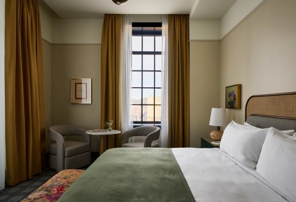 A neatly made bed with a green throw invites rest in a hotel room featuring a tall window with city views, two armchairs, and artwork on cream walls.