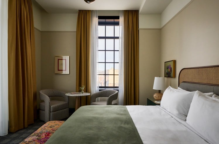 A neatly made bed with a green throw invites rest in a hotel room featuring a tall window with city views, two armchairs, and artwork on cream walls.