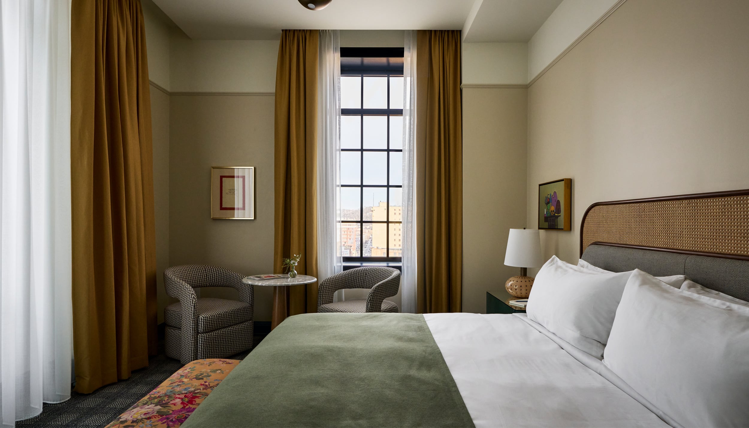 A neatly made bed with a green throw invites rest in a hotel room featuring a tall window with city views, two armchairs, and artwork on cream walls.