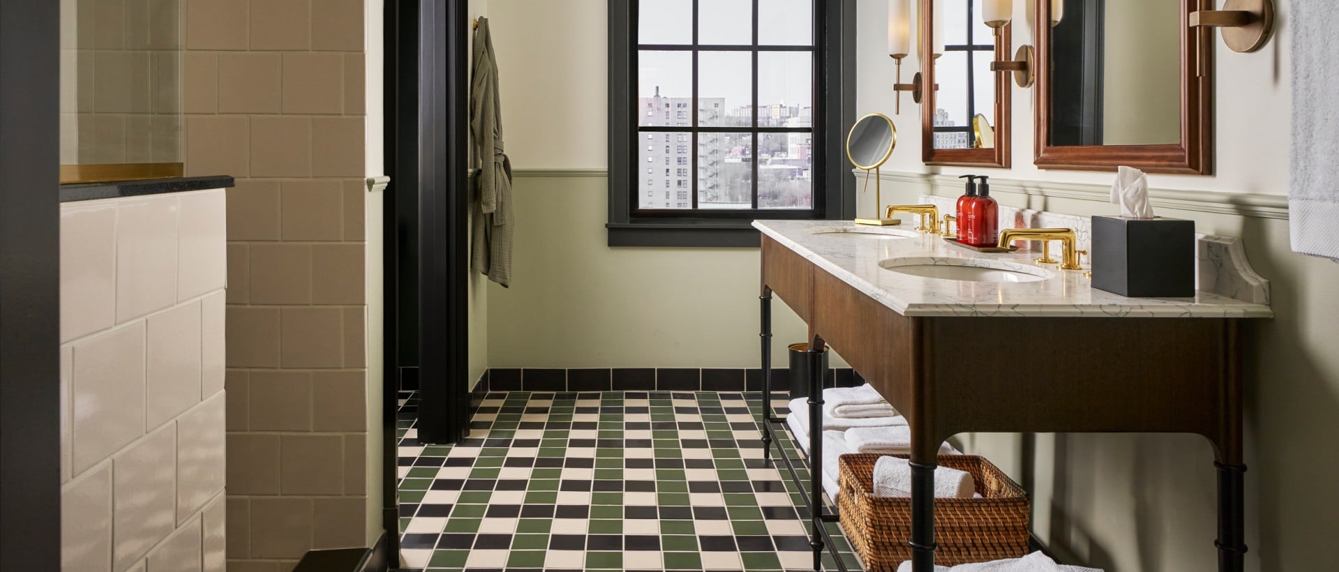 Marble vanity with gold faucets, mirrors, and two red "LITY" bottles. The modern bathroom has a green/black checkered floor and a window showing city buildings.