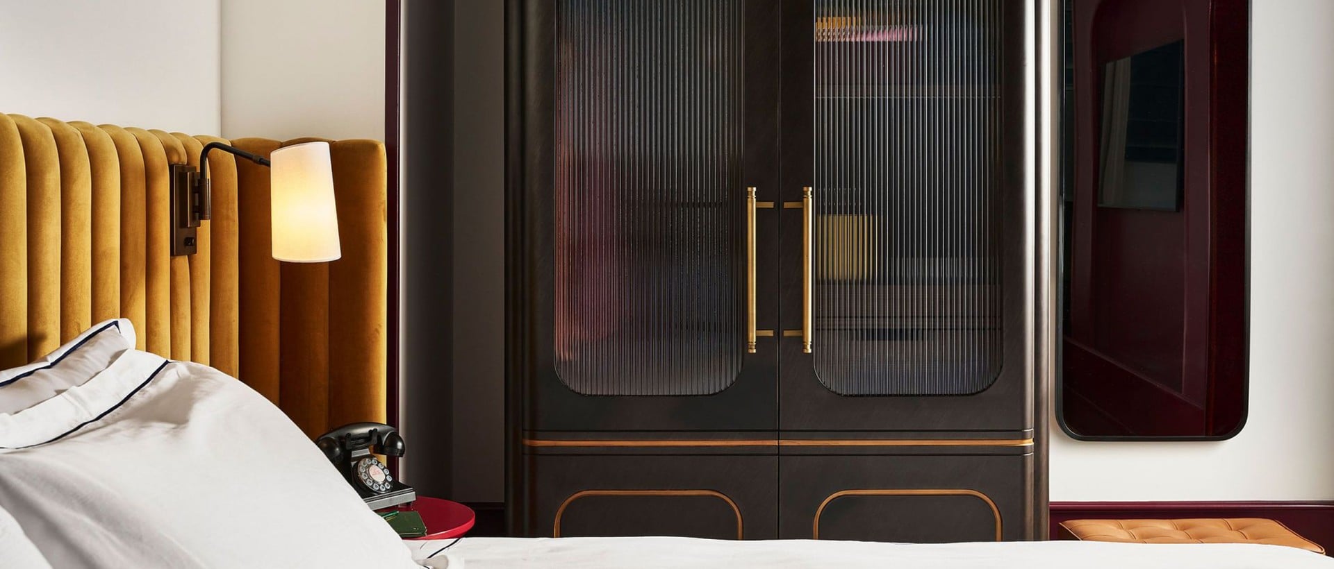 Mustard yellow headboard and dark fluted-glass wardrobe frame a bed. A wall lamp glows in a stylish bedroom featuring a vintage phone and mirror.