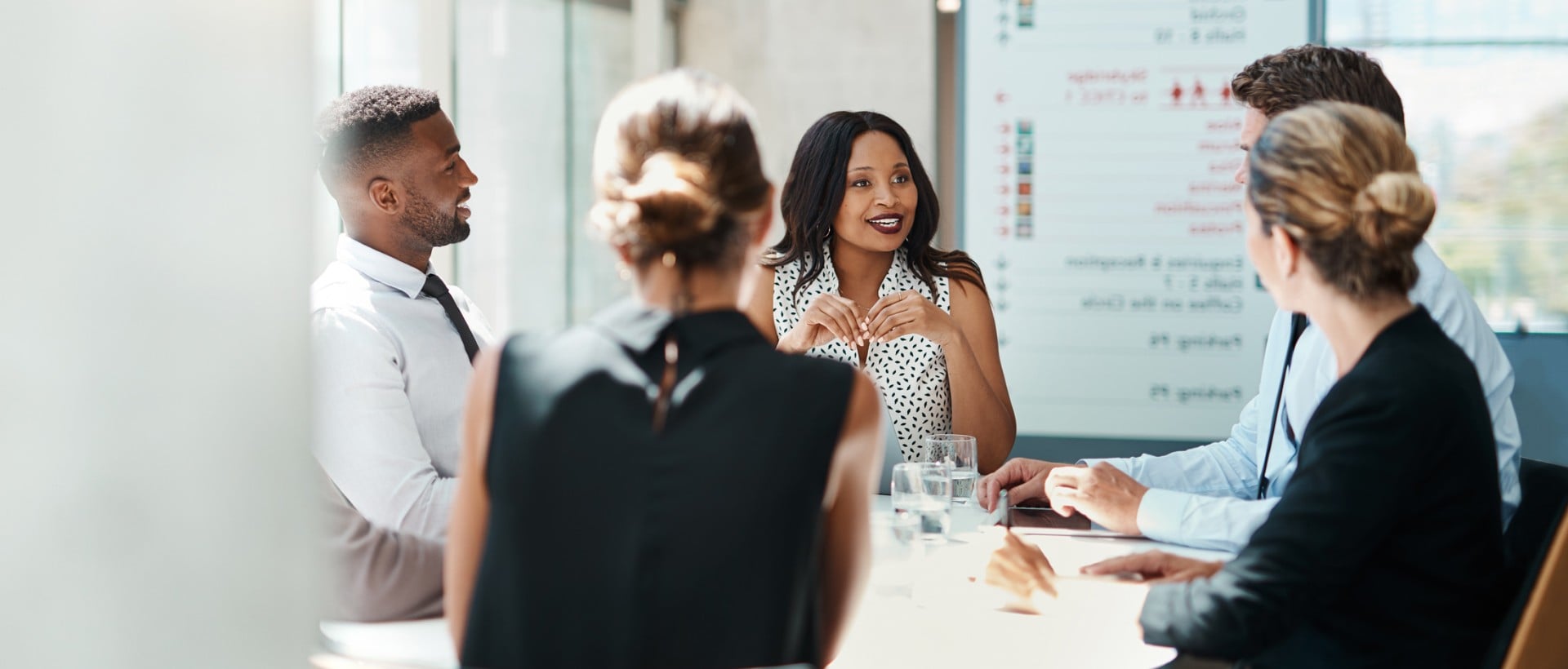 Five professionals sit around a white table, a woman speaking during a bright office meeting. A whiteboard displays blurry text, a large '3', an 'S', and four red star symbols.
