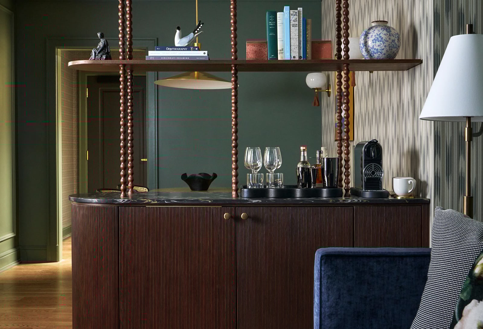 A dark fluted wood bar cabinet with a marble top displays barware and a coffee machine, beneath a suspended shelf holding books titled "ORNITHOGRAPHY", "CINCINNATI DREAMING", "THE WORK", "ADAM", "ART", "NOVELS", against green and striped walls.