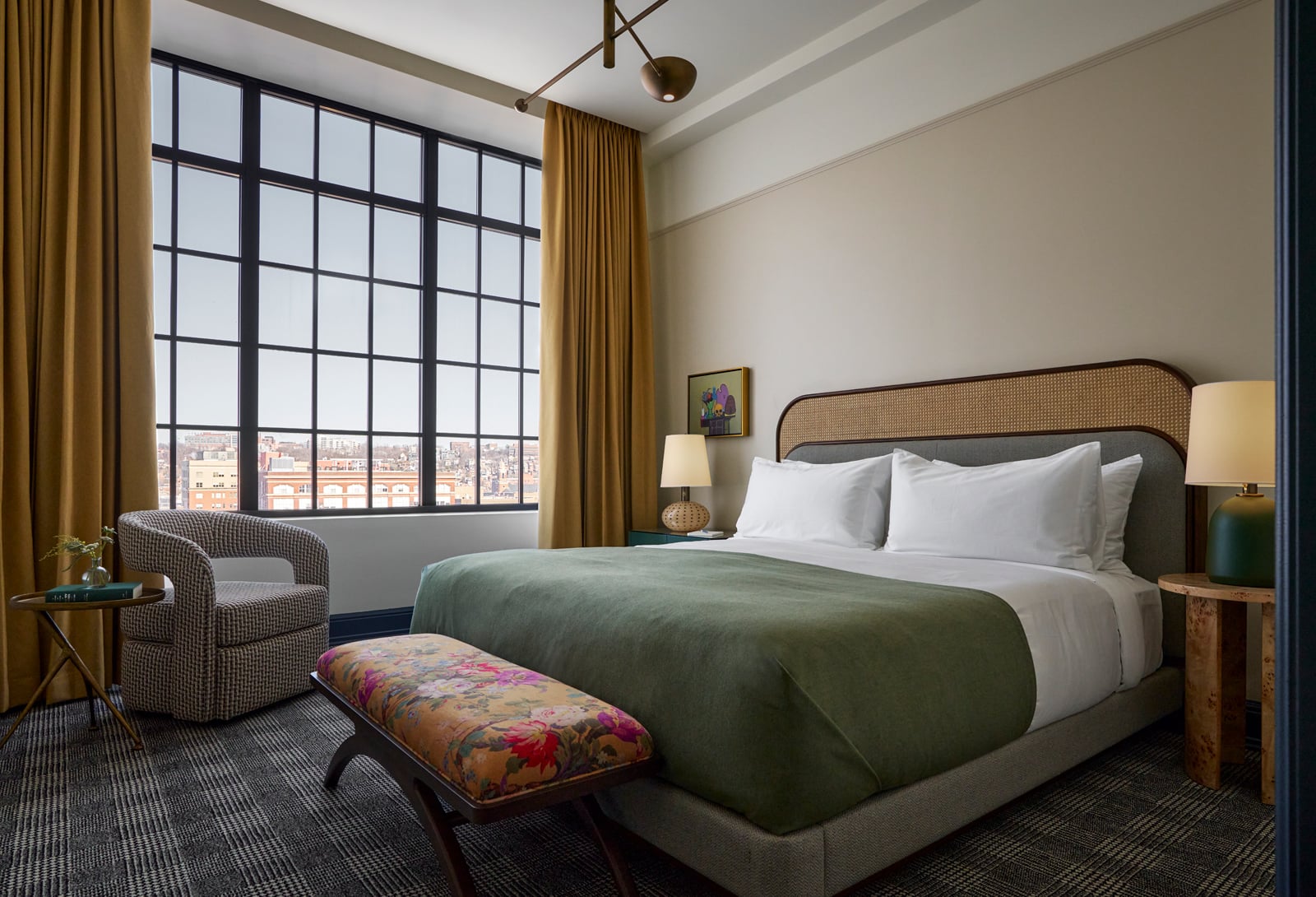 A made bed with green duvet and a floral bench at its foot is the focus of a hotel room. A large gridded window overlooks a city.