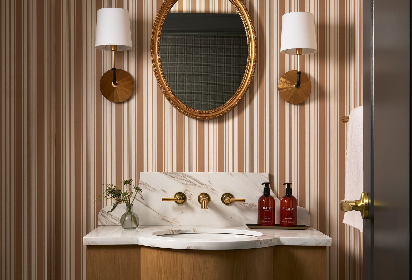 A bathroom vanity, featuring a marble counter, oval sink, and brass wall faucet, sits below a gold-framed mirror and sconces on a striped wall. Two bottles read: "FIDELITY HOTEL hand soap sweet fig + rosemary 500ml e 16.9 fl.oz.", "FIDELITY HOTEL body lotion sweet fig + rosemary 500ml e 16.9 fl.oz.".