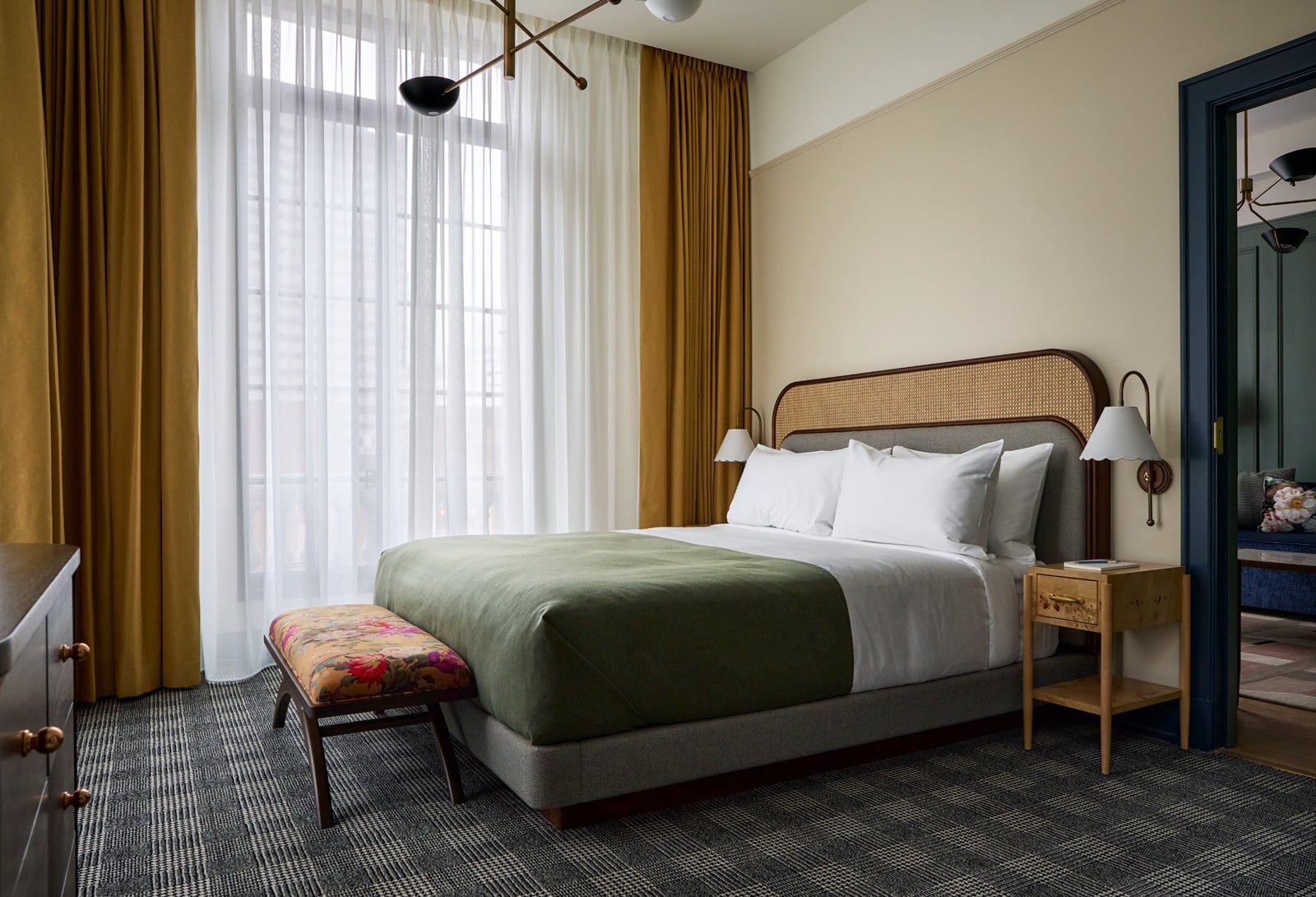 A bed with a cane headboard, white sheets, and a green duvet, sits in a stylish room with plaid carpet, a floral bench, and gold window curtains.