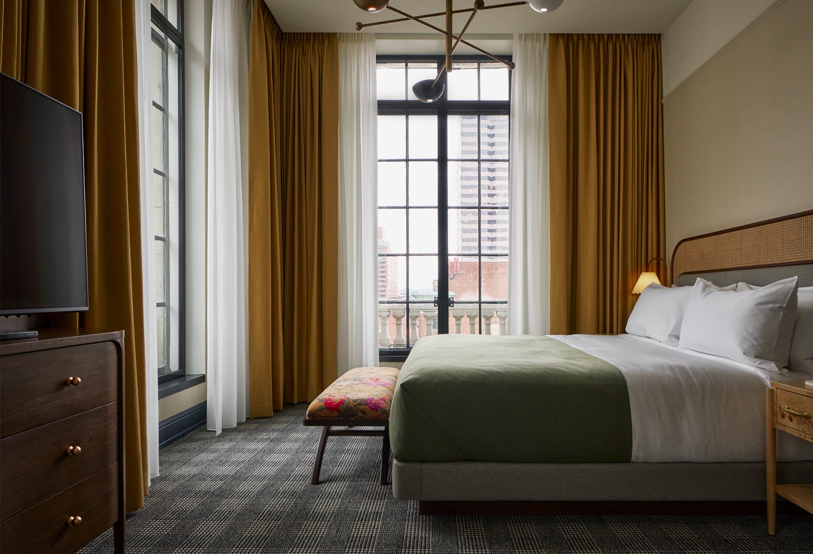 A modern hotel bedroom with a large bed, featuring a green throw, faces tall windows with city views. Gold curtains, a TV on a dresser, and a floral bench furnish the room.