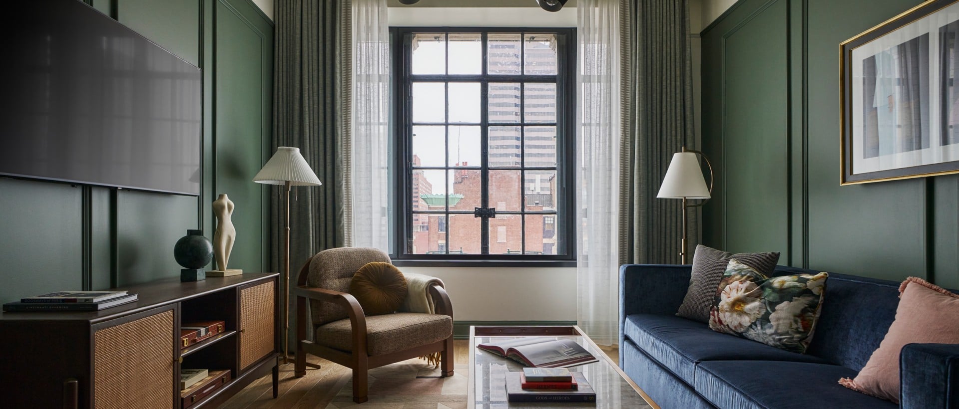 A modern living room with a blue velvet sofa, brown armchair, and glass coffee table. A TV is mounted on a dark green paneled wall, facing a city view window. Text found: HERMÈS, GODS OF HEROES, DOORWAYS OF PARIS, CINCINNATI DREAMING.
