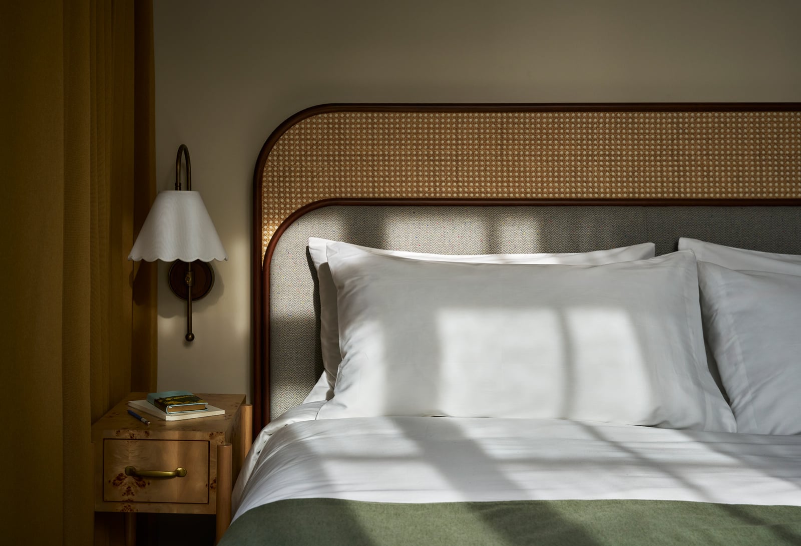 A neatly made bed with white pillows, a green blanket, and a rattan/fabric headboard receives sunlit patterns. A wall sconce and wooden nightstand sit next to it.