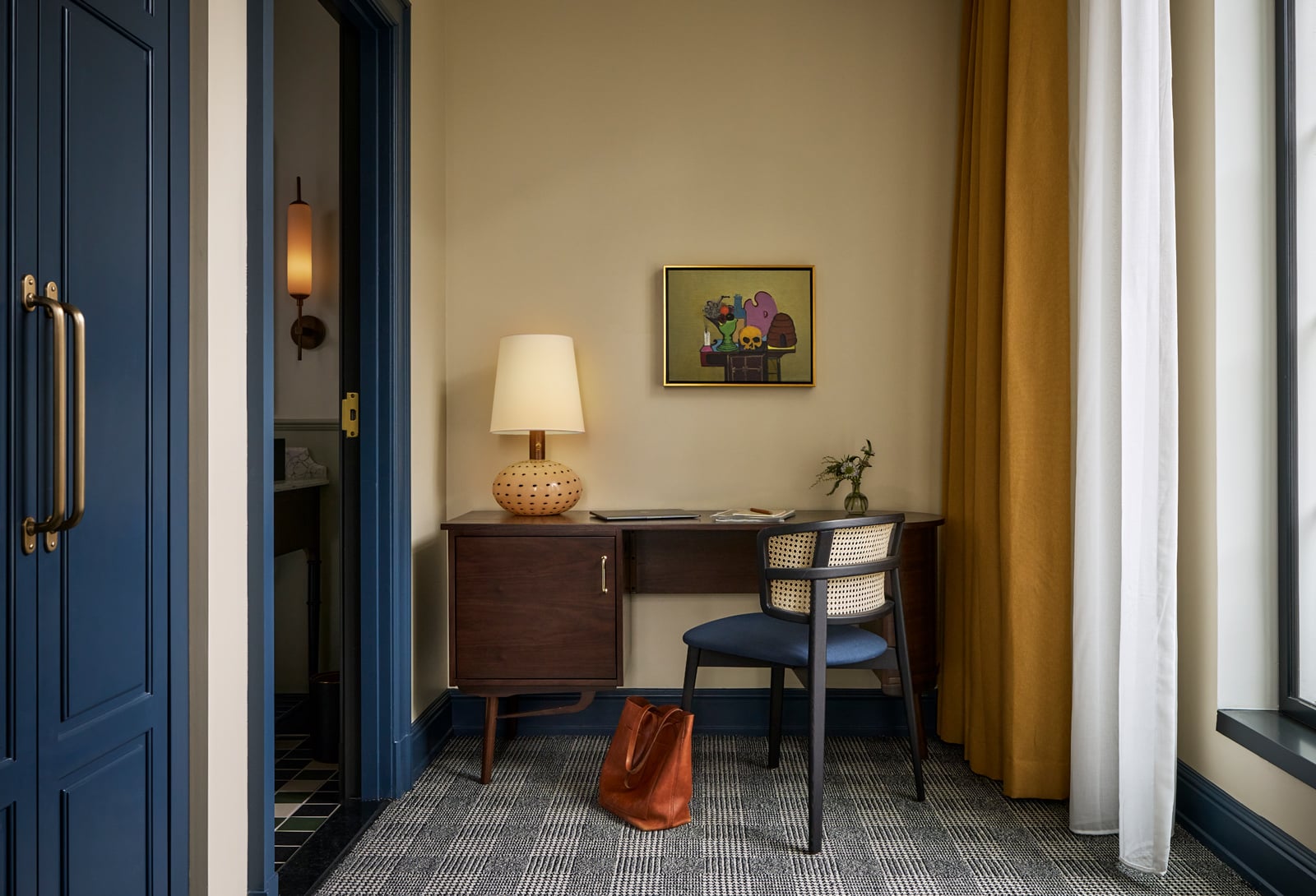 A dark wood desk with a patterned lamp and cane chair furnishes a neutral-walled room with plaid carpet. An abstract painting hangs above, flanked by a blue doorway and window.