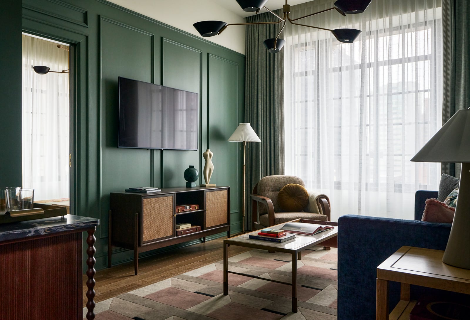 A modern living space showcases a wall-mounted TV above a rattan cabinet set against a dark green paneled wall, with a large, sheer-curtained window, stylish armchair, and geometric rug.