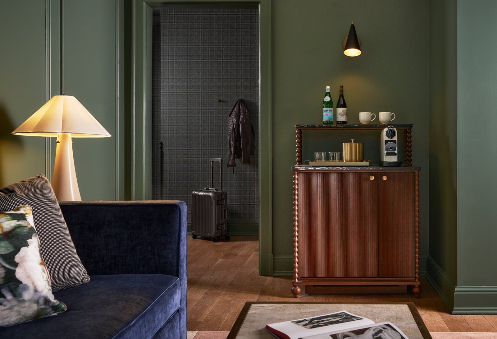 Dark wood mini-bar cabinet with PELLEGRINO, coffee machine, cups, against a dark green wall. Blue sofa foreground; suitcase and jacket seen through doorway.