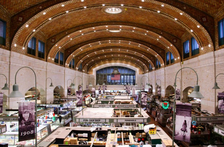Market hall interior, with stalls displaying goods under a vaulted brick ceiling. It is a grand, historic space with an American flag and clock at its far end.