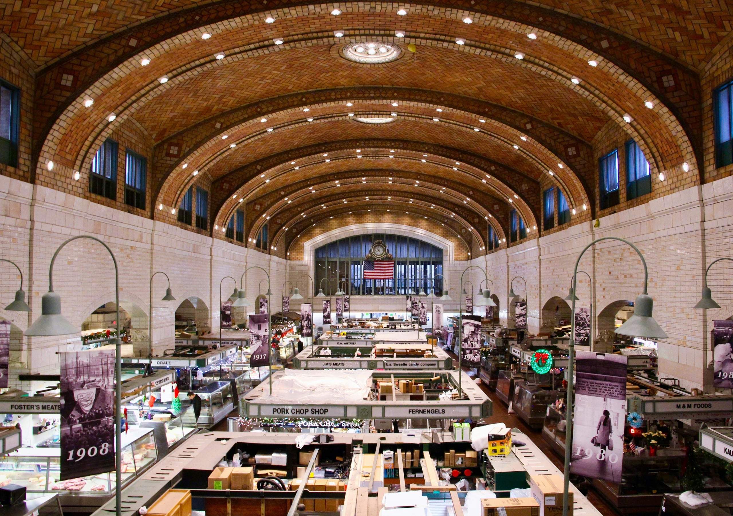 Market hall interior, with stalls displaying goods under a vaulted brick ceiling. It is a grand, historic space with an American flag and clock at its far end.