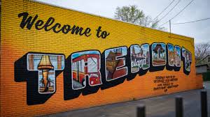 A large mural on a yellow brick wall displays "Welcome to TREMONT" in postcard-style letters, each depicting a local scene. Text: Welcome to TREMONT DESIGNED & PAINTED BY AARON & RACHAEL DANCEY AND JOHN HACKER DANCEY STUDIOS. TREMONTARTS.ORG