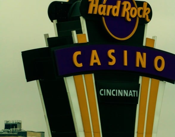 A tall, multi-tiered "Hard Rock CASINO CINCINNATI" sign features gold lettering on purple, with black, white, and gold panels, against a pale, hazy sky and distant buildings.