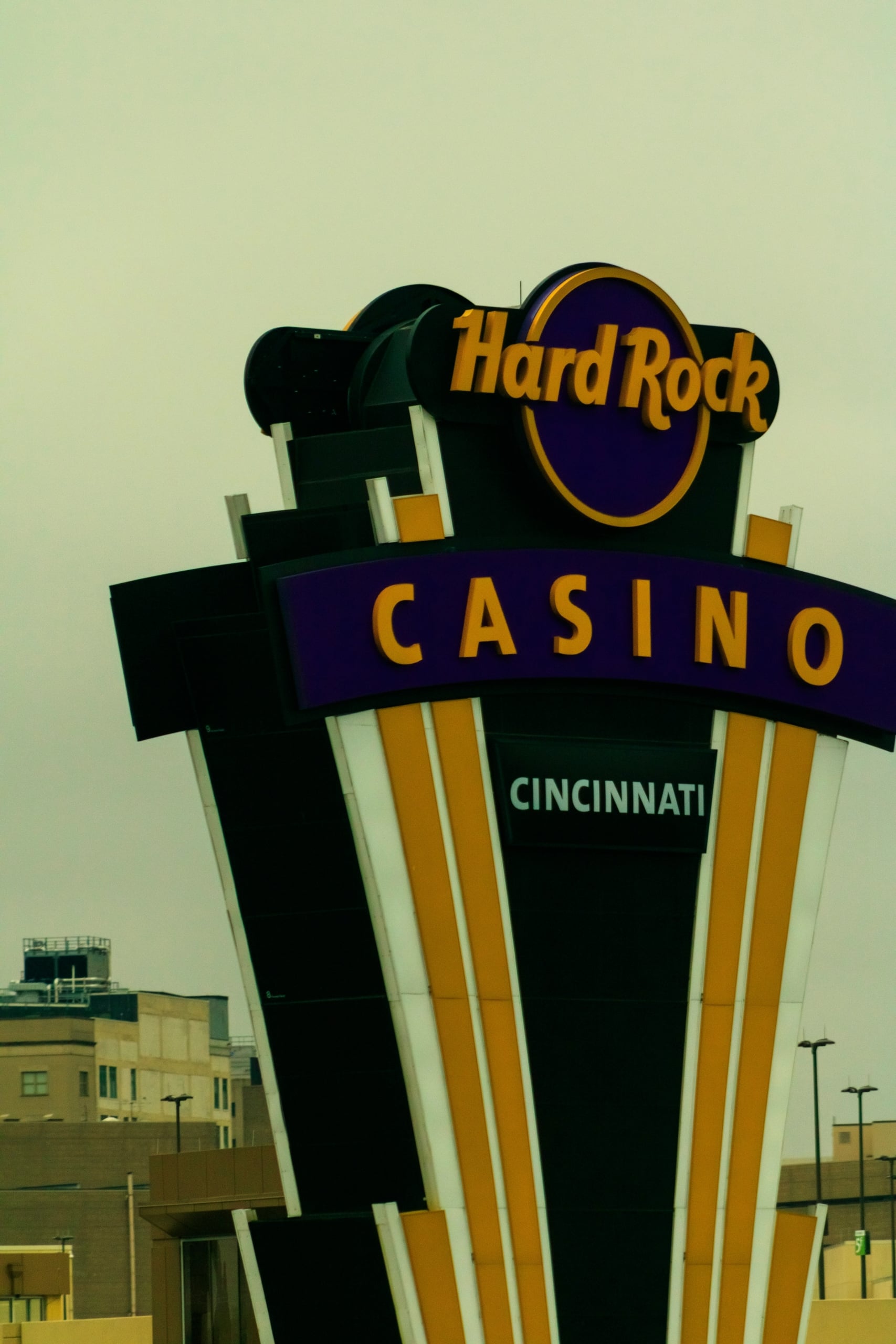 A tall, multi-tiered "Hard Rock CASINO CINCINNATI" sign features gold lettering on purple, with black, white, and gold panels, against a pale, hazy sky and distant buildings.