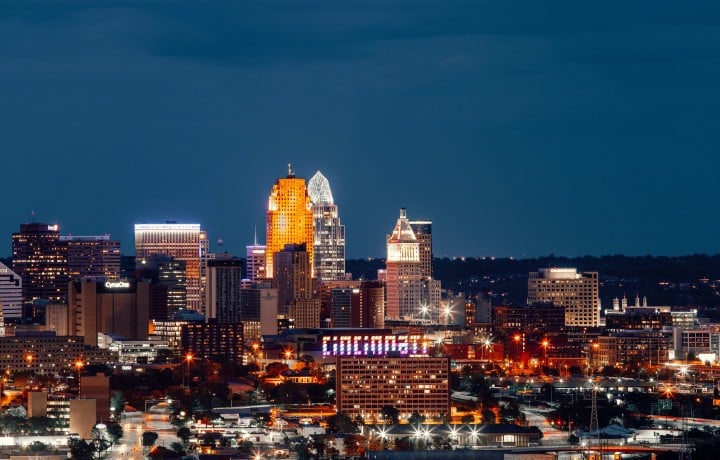 A city skyline glows at night, showcasing illuminated buildings and streetlights. Visible text includes: "Fifth Third", "Macy's", "P&G", "CyrusOne", "MILLENNIUM HOTEL", "Burke", "Delta Energy Center", "Cincinnati.com", and a large "CINCINNATI" sign. All against a dark blue evening sky.