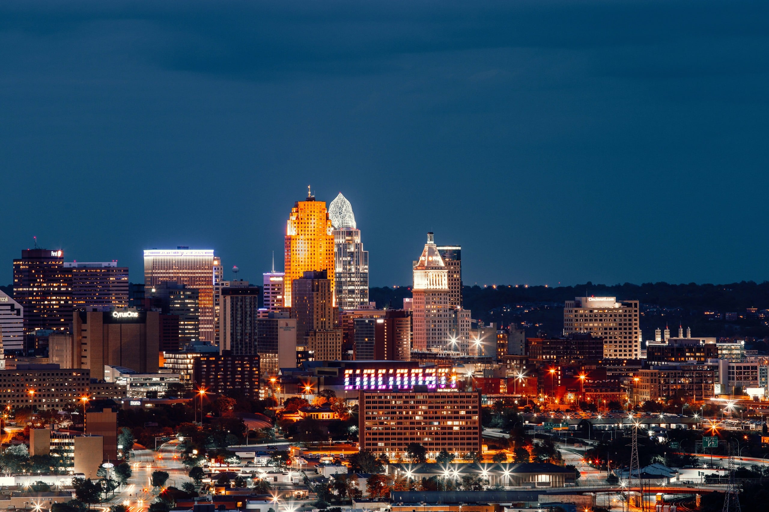 A city skyline glows at night, showcasing illuminated buildings and streetlights. Visible text includes: "Fifth Third", "Macy's", "P&G", "CyrusOne", "MILLENNIUM HOTEL", "Burke", "Delta Energy Center", "Cincinnati.com", and a large "CINCINNATI" sign. All against a dark blue evening sky.