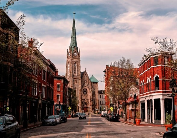 A Gothic church with a green spire and clock dominates a city street lined with red brick buildings and parked cars, under a cloudy sky. Text includes: CLAYTON, WOODBURN, FREE LOCAL EL FOCO.