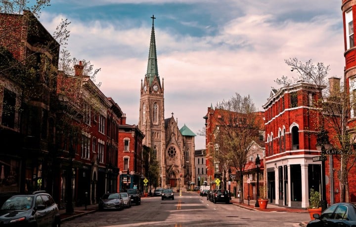 A Gothic church with a green spire and clock dominates a city street lined with red brick buildings and parked cars, under a cloudy sky. Text includes: CLAYTON, WOODBURN, FREE LOCAL EL FOCO.