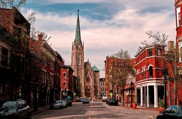 A Gothic church with a green spire and clock dominates a city street lined with red brick buildings and parked cars, under a cloudy sky. Text includes: CLAYTON, WOODBURN, FREE LOCAL EL FOCO.