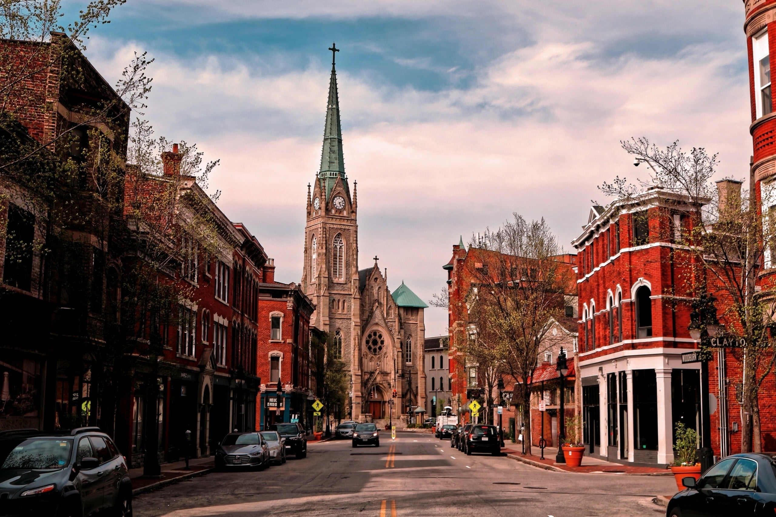 A Gothic church with a green spire and clock dominates a city street lined with red brick buildings and parked cars, under a cloudy sky. Text includes: CLAYTON, WOODBURN, FREE LOCAL EL FOCO.