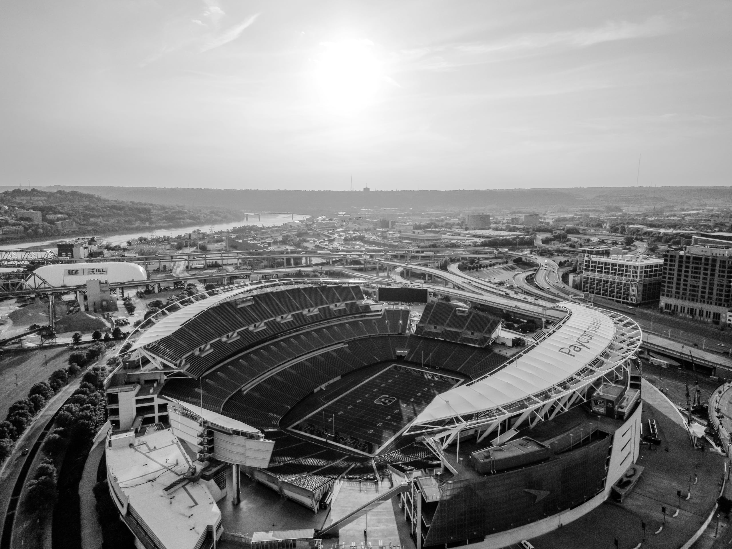 Aerial, monochromatic view of a large, empty football stadium (Paycor Stadium, BENGALS, GEICO, BETFRED, NFL) in a sprawling city with highways, a river, and distant hills under a hazy sky.