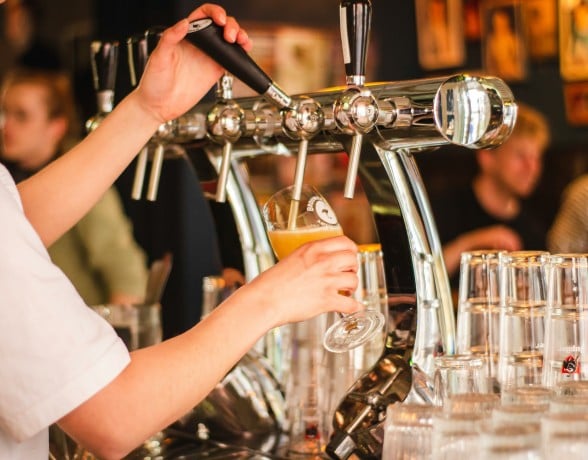 Bartender's hands operate a "BROUW" tap, pouring a "BROUWERIJ AMSTERDAM" beer into a glass. Stacked "Jupiler" glasses sit on the bar, blurred patrons in the background.