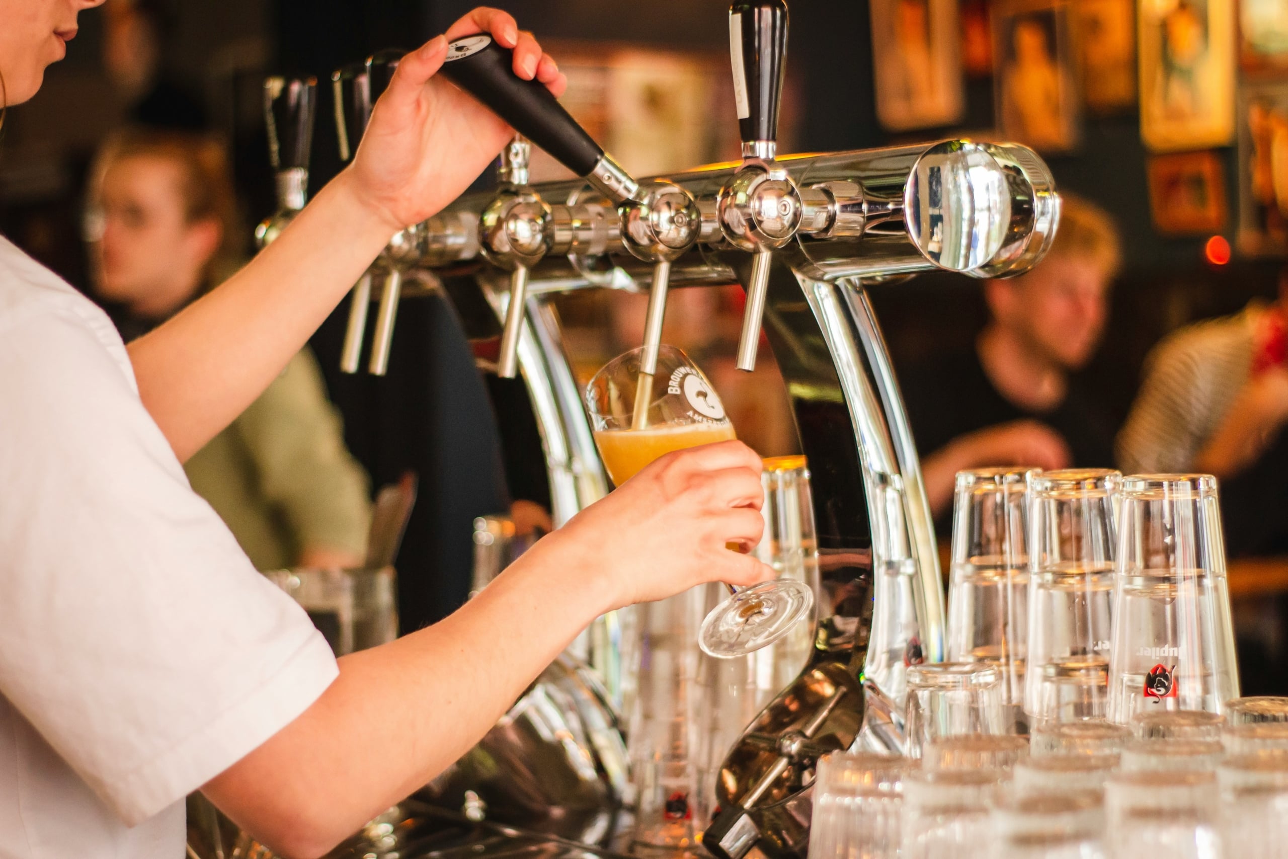 Bartender's hands operate a "BROUW" tap, pouring a "BROUWERIJ AMSTERDAM" beer into a glass. Stacked "Jupiler" glasses sit on the bar, blurred patrons in the background.